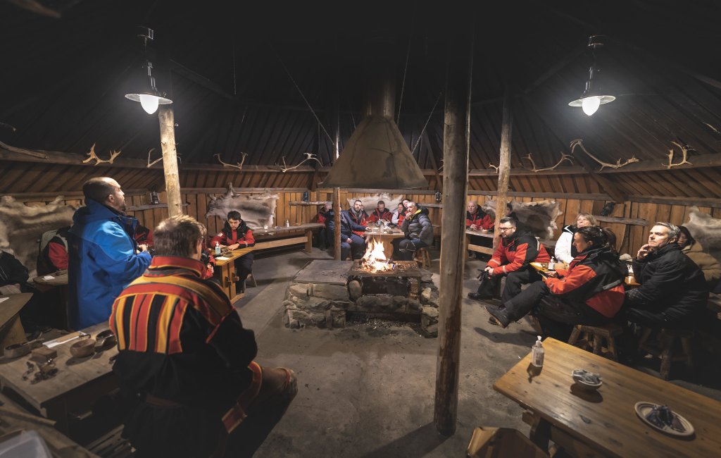 Group of people by a fireplace in a hut.